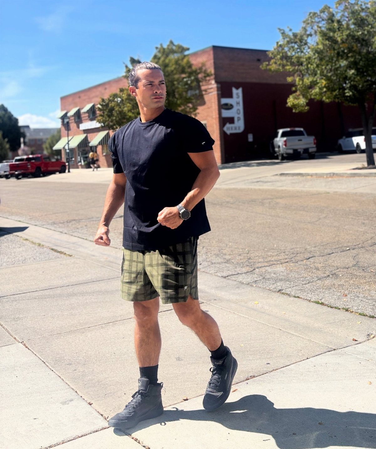 Man walking on sidewalk wearing black t-shirt and olive green plaid shorts