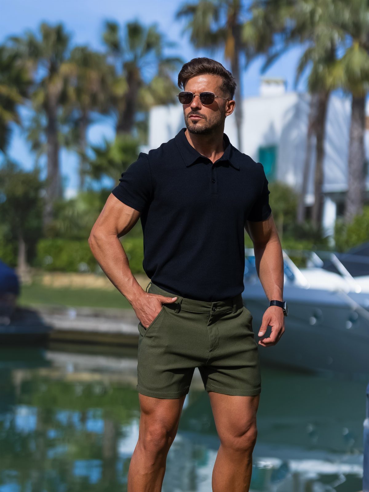 Man in black polo and olive green chino shorts standing by the water.
