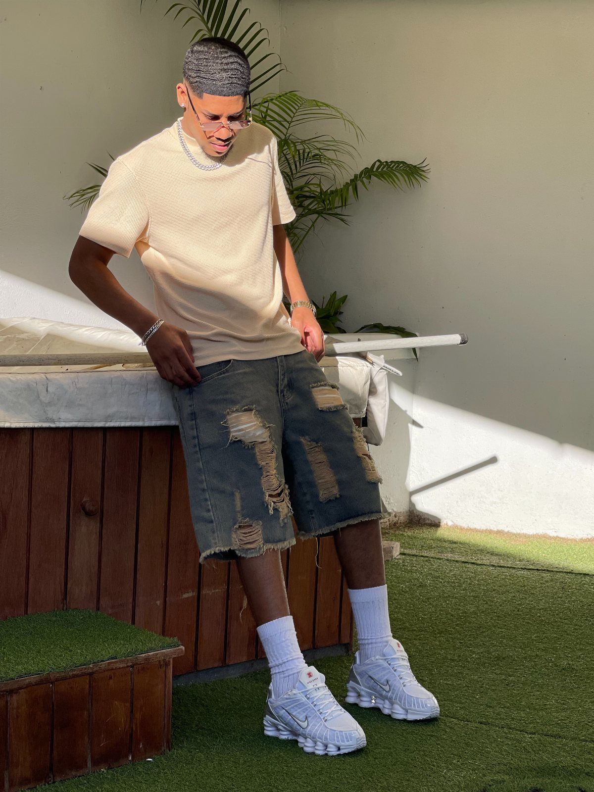 Man wearing distressed denim shorts, cream t-shirt, and white sneakers on green turf.