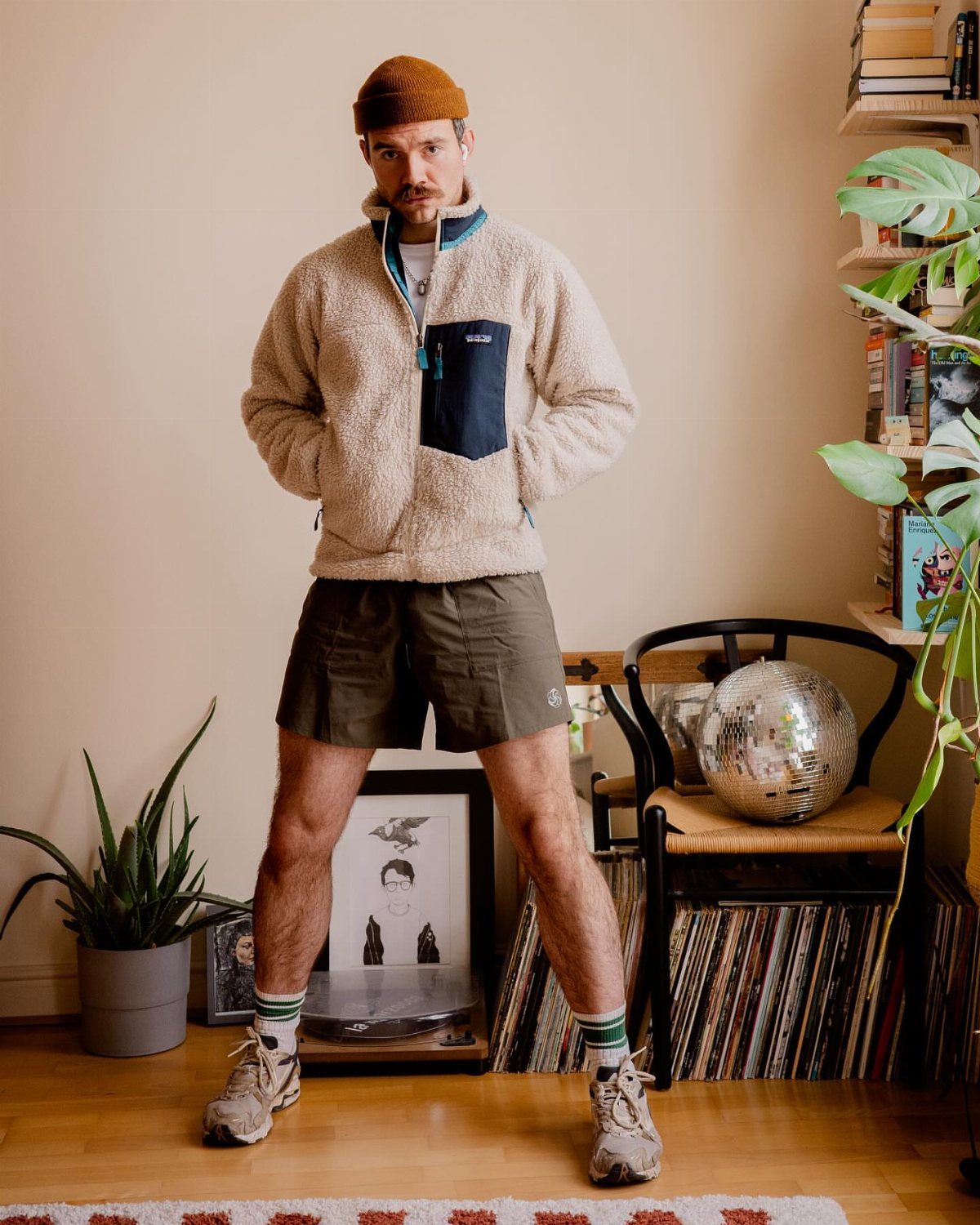 Man wearing olive green hiking shorts, beige fleece jacket, and hiking boots indoors.
