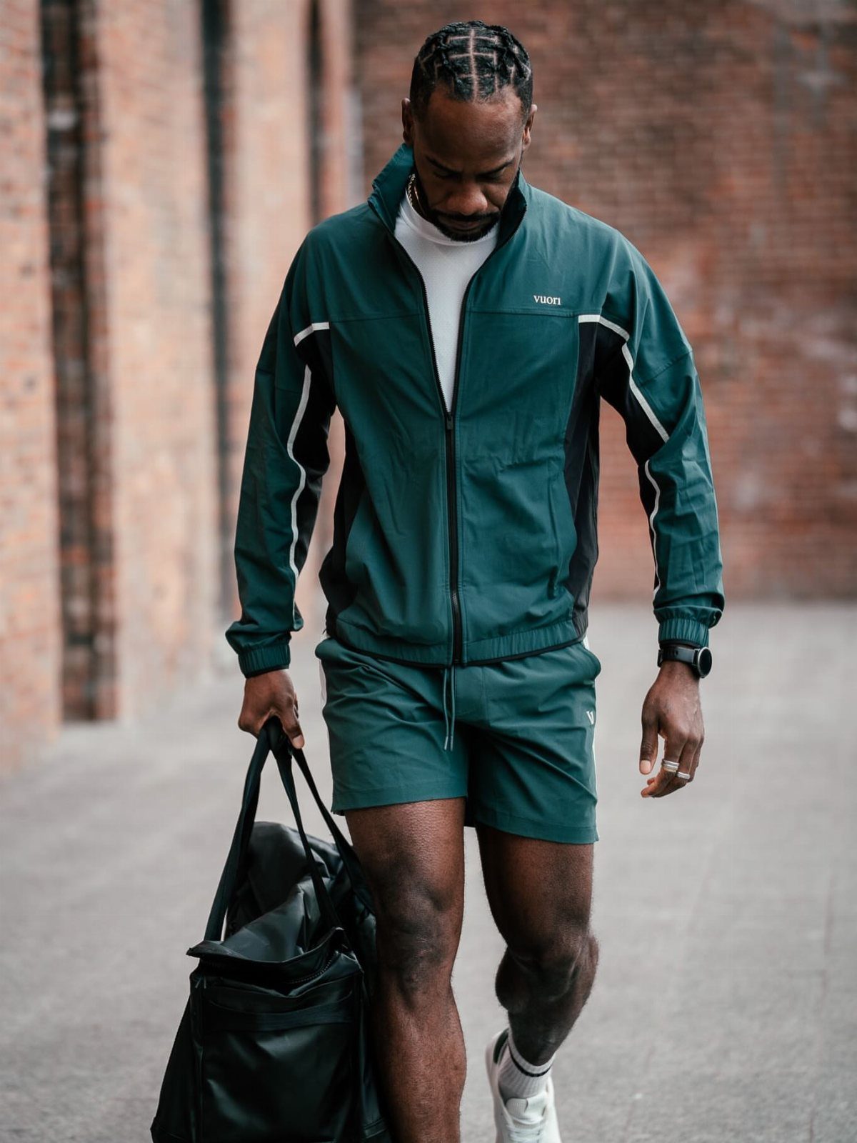 Man in dark green athletic jacket and shorts carrying a black gym bag.