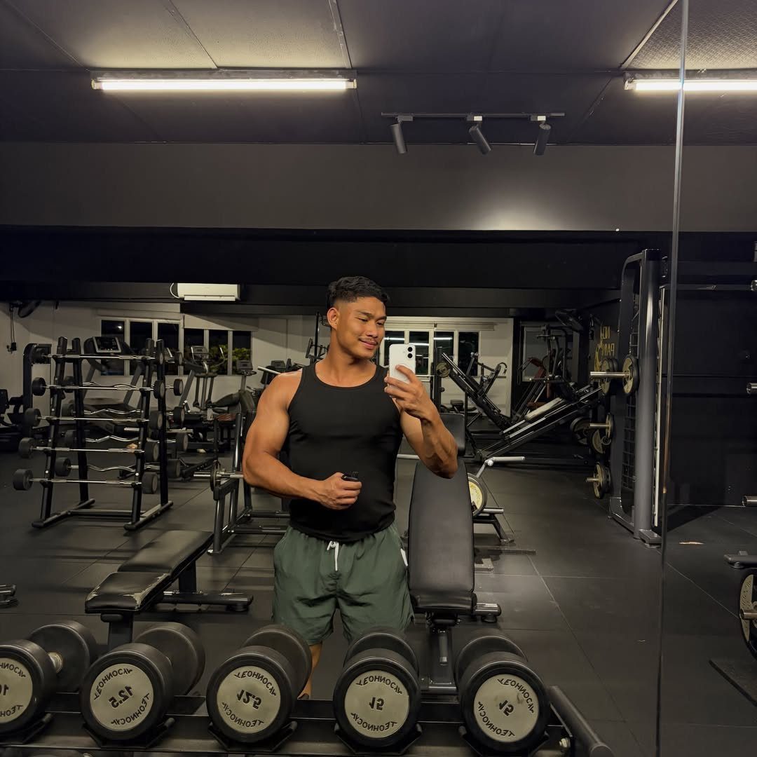 Muscular man in black sleeveless tank top and green shorts posing in gym mirror.