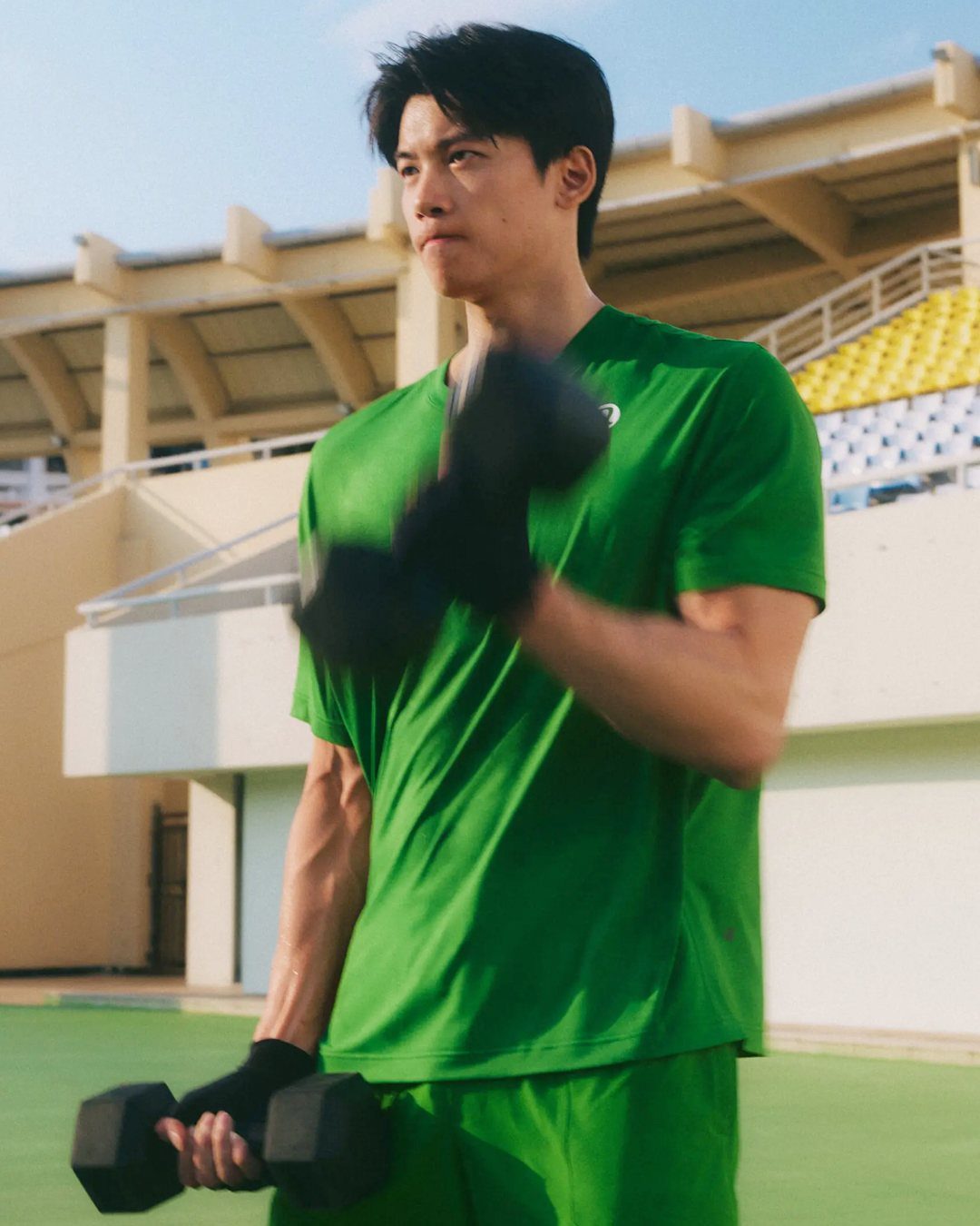 Man in green athletic wear lifting dumbbells at an outdoor stadium.