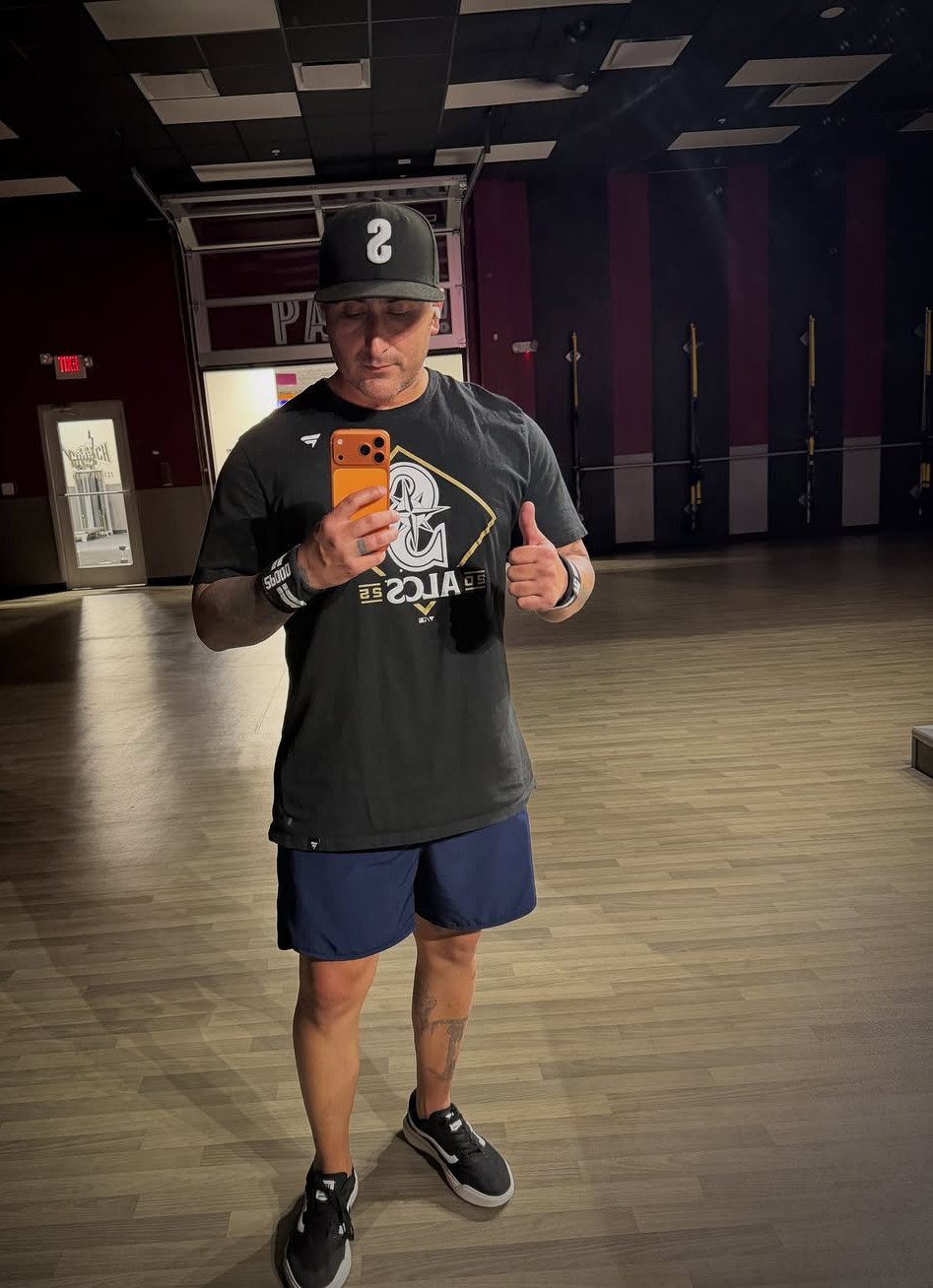 Man in black t-shirt and blue athletic shorts taking a gym mirror selfie.