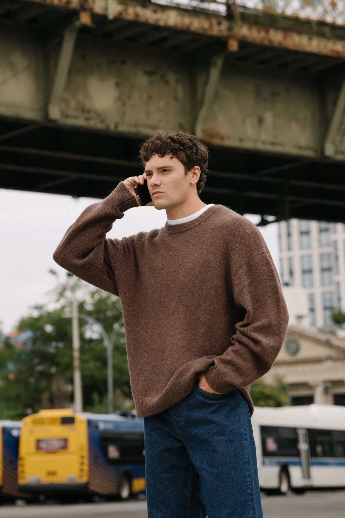 Man wearing a brown oversized knit sweater and blue jeans talking on a phone