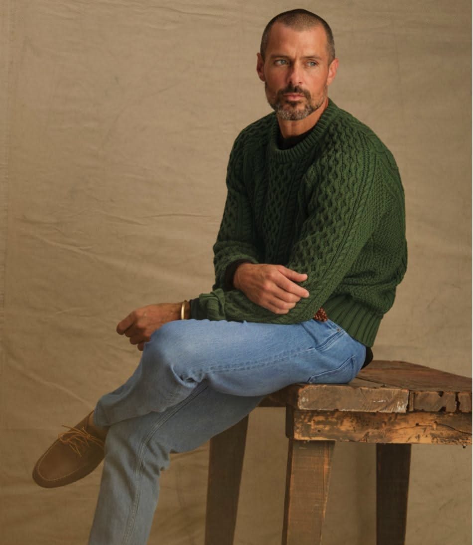Man wearing olive green cable-knit sweater and light blue jeans sitting on wooden stool