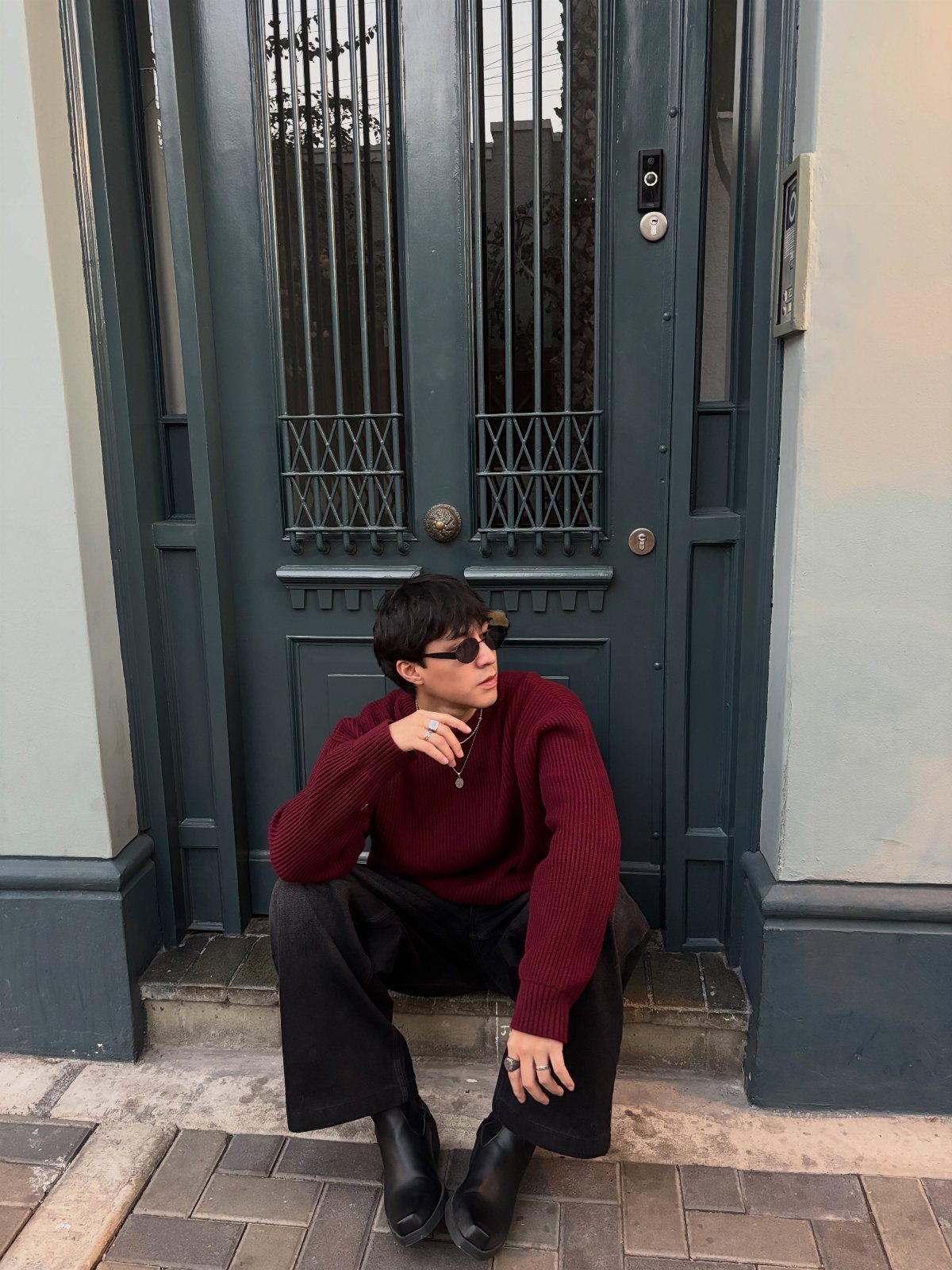 Person in red oversized sweater and black jeans sitting before a teal door.