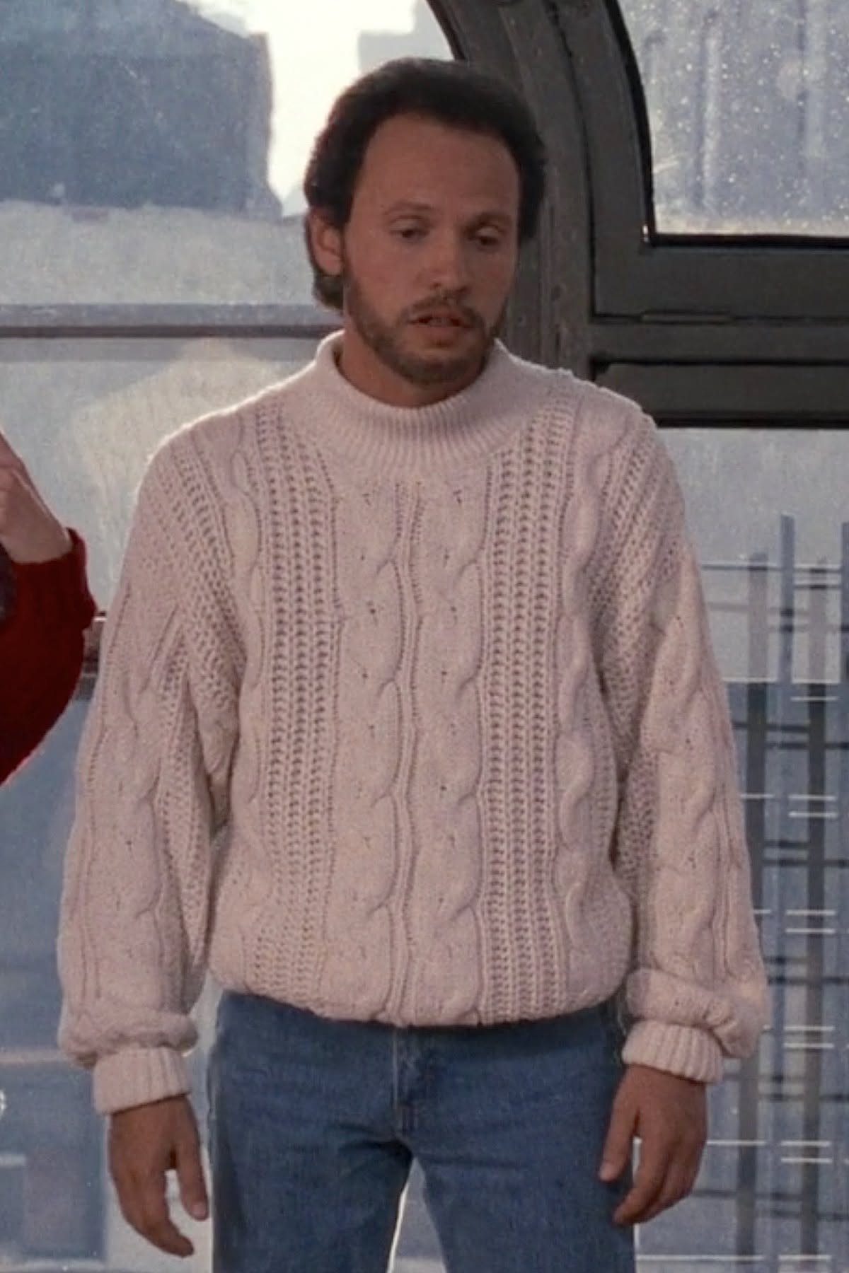 Man wearing a white cable-knit sweater and blue jeans standing by a window.