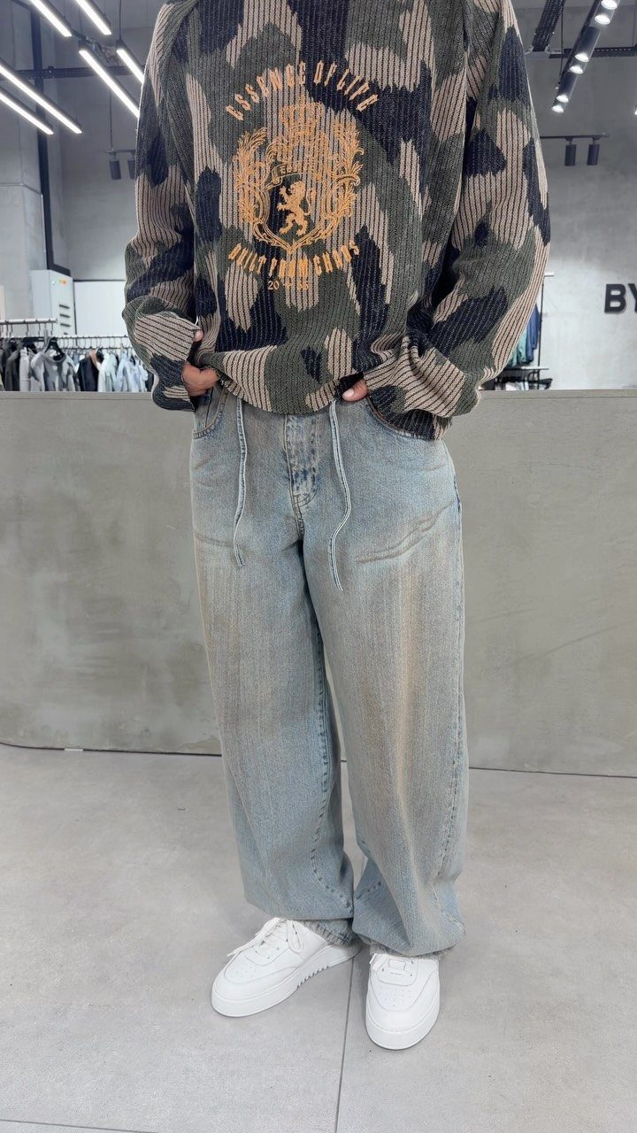 Person wearing a camouflage sweater, wide-leg light blue jeans, and white sneakers.