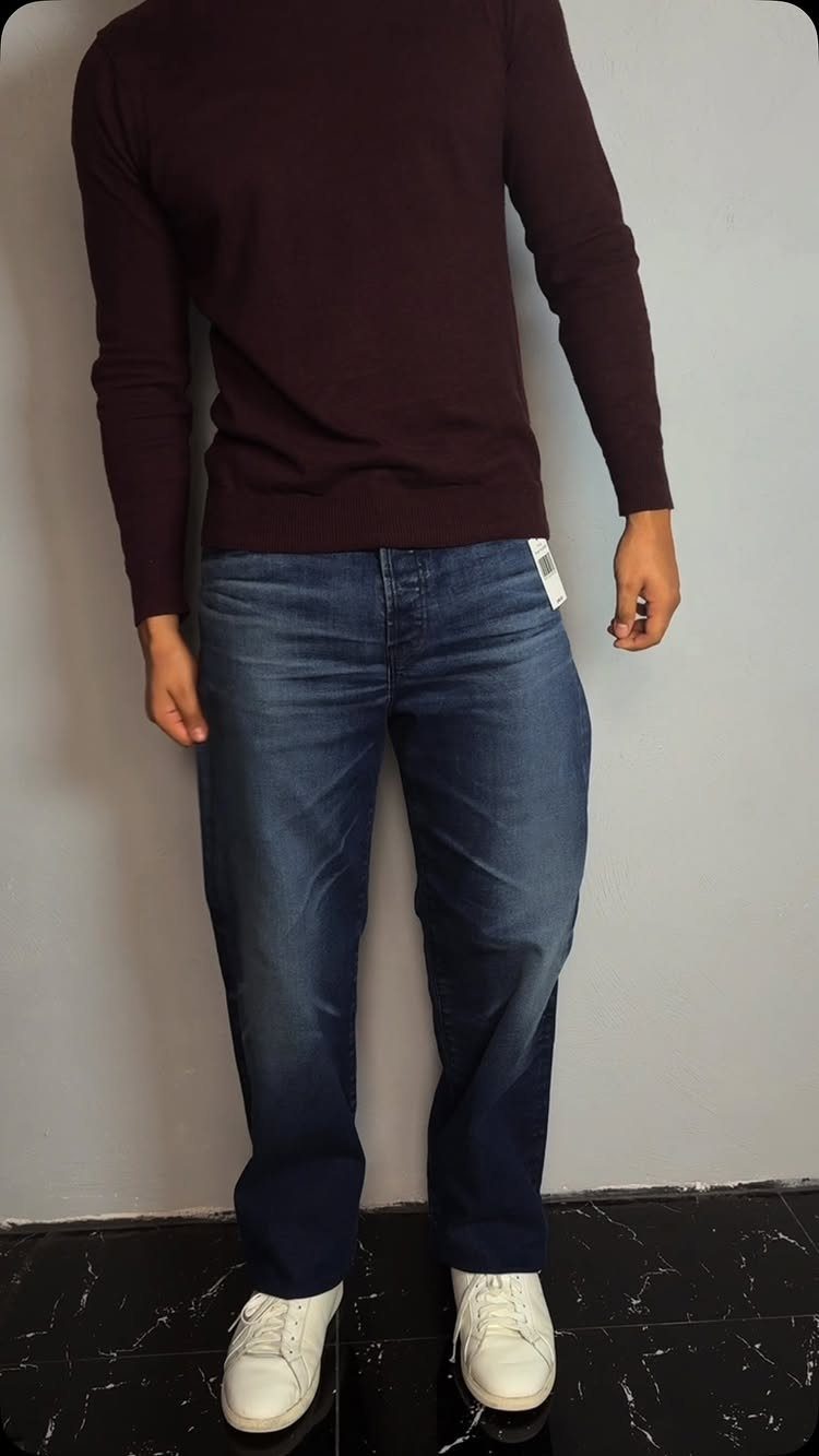 Person wearing a burgundy sweater, blue jeans, and white sneakers against a grey wall.