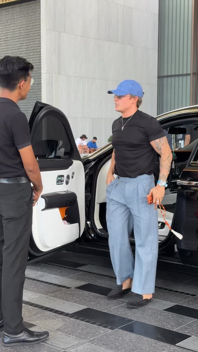 Man in black t-shirt and light blue wide-leg jeans standing by a car.