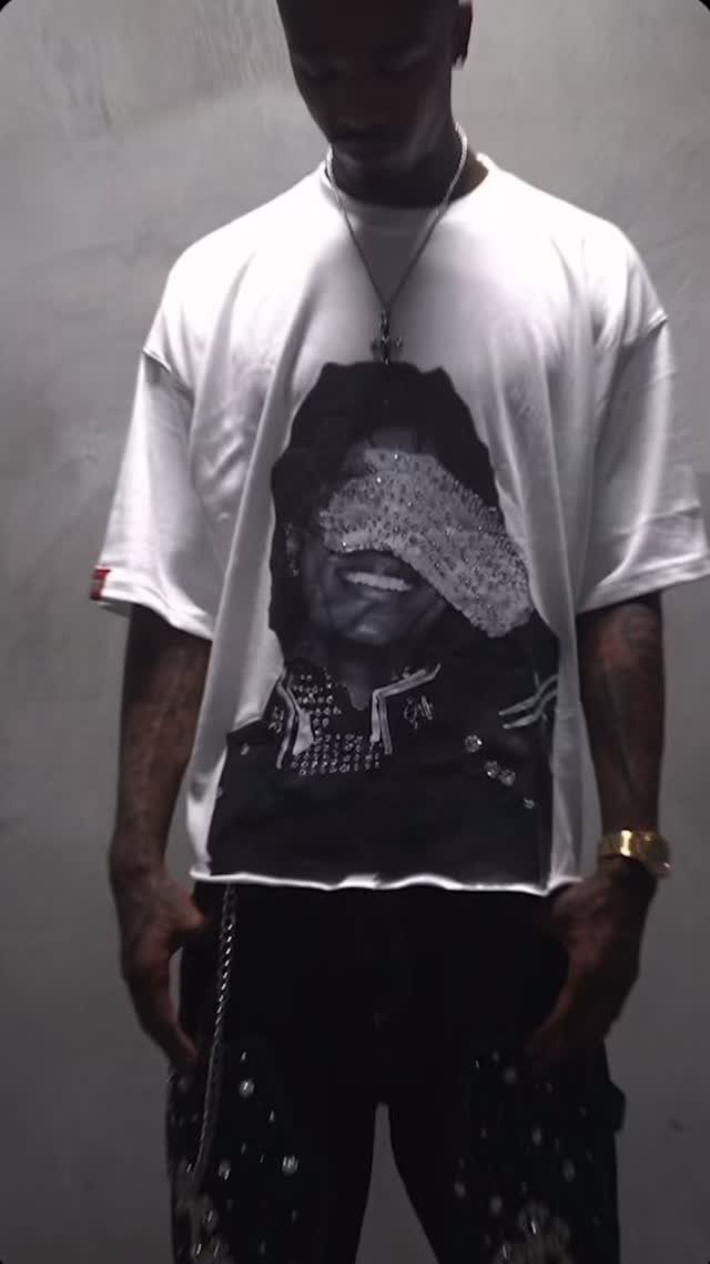 Person wearing a white graphic t-shirt and black embellished jeans against a grey wall.