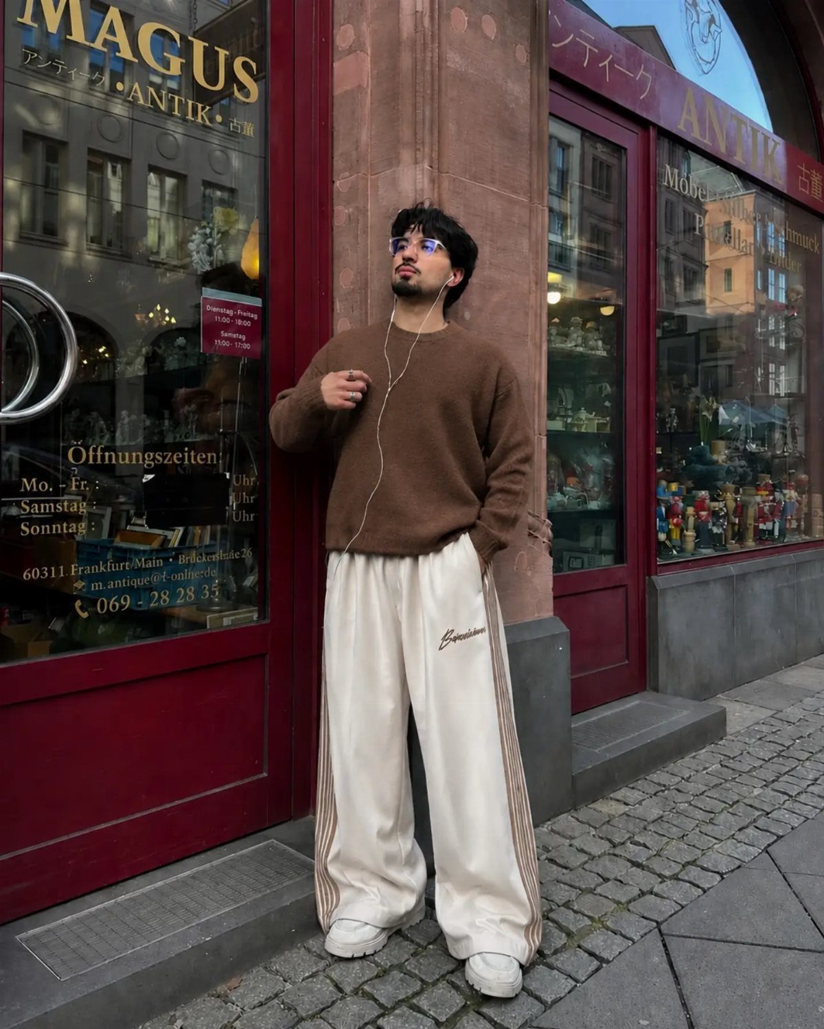 Man in brown sweater and wide-leg cream track pants standing on city street.