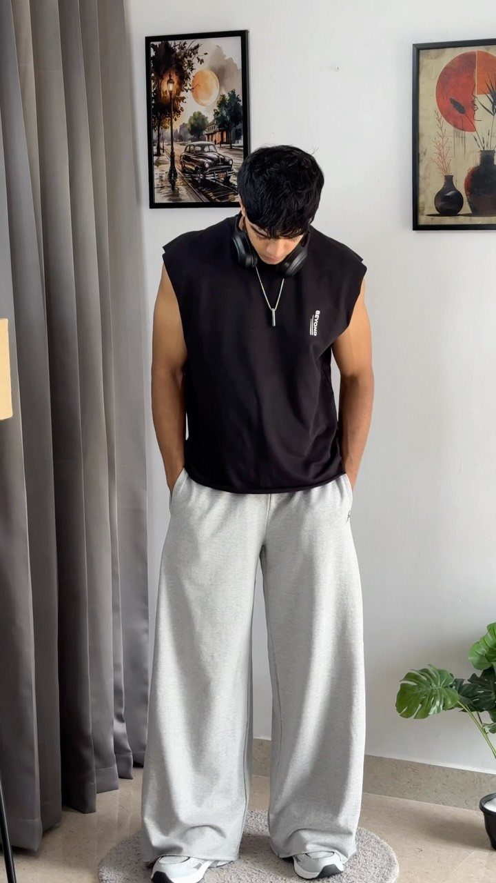 Man wearing a black sleeveless top and wide-leg grey track pants indoors.
