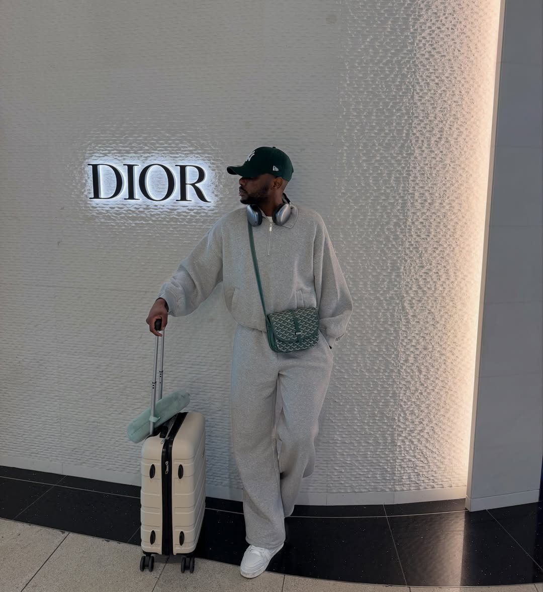 Man in grey track suit with suitcase standing before a Dior store wall.