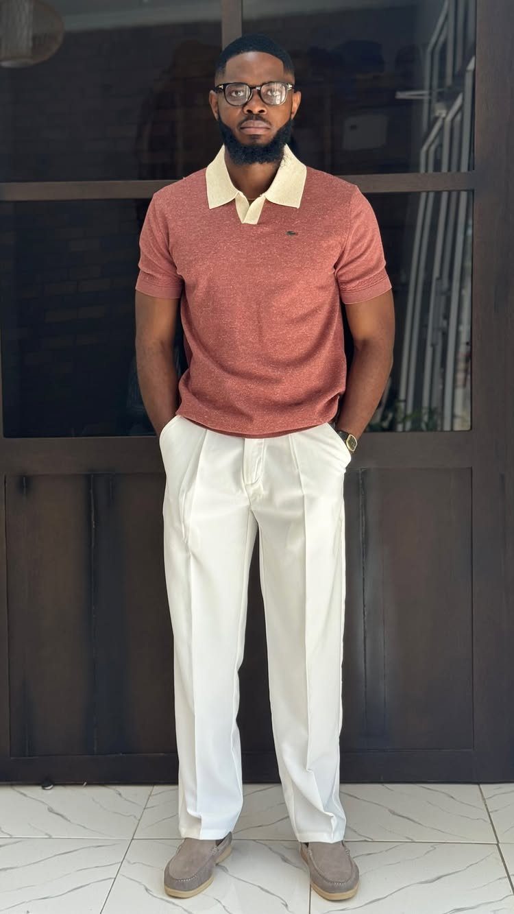 Man wearing a terracotta polo shirt and white pleated trousers standing indoors.