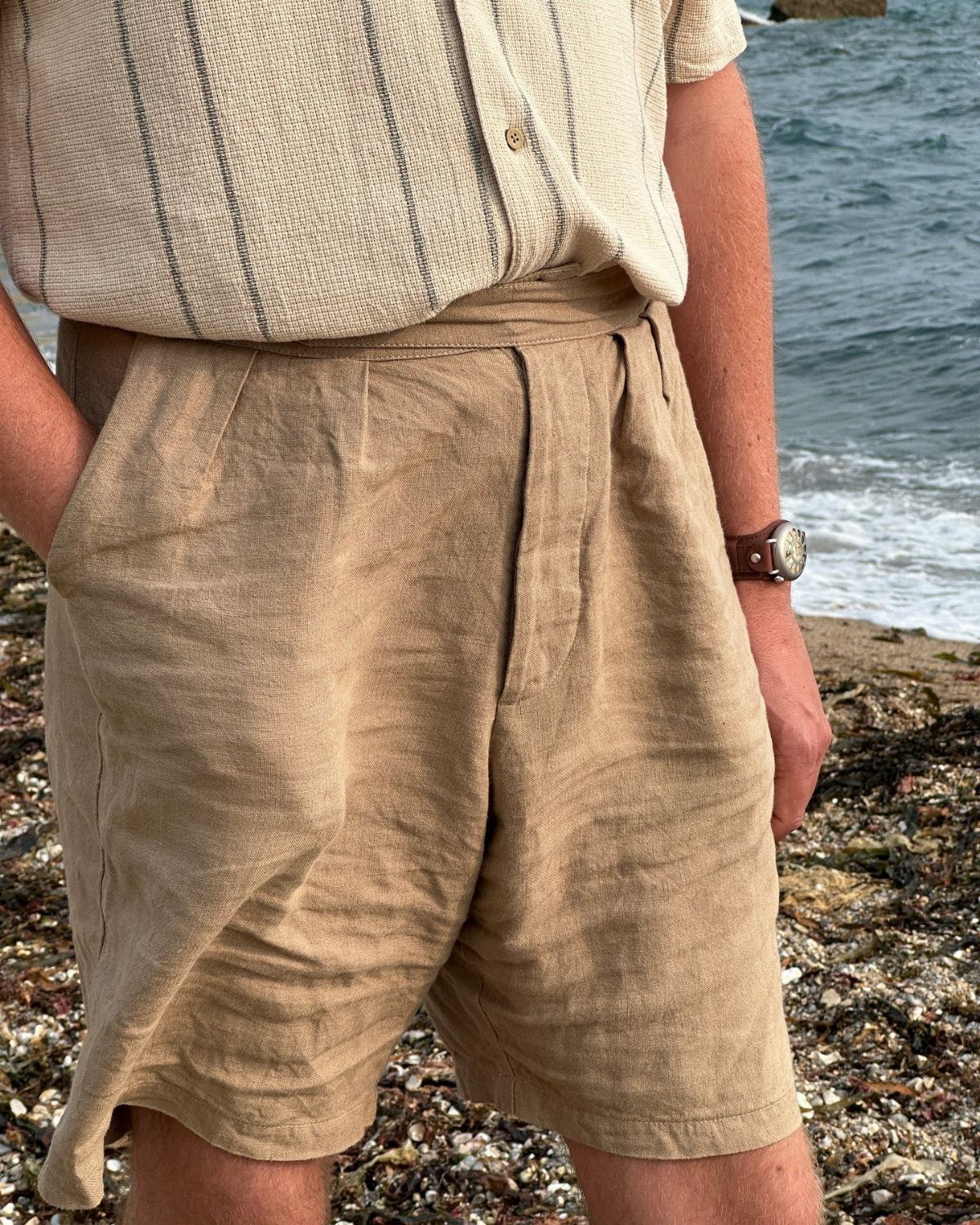 Tan pleated linen shorts worn by a person on a pebbly beach.