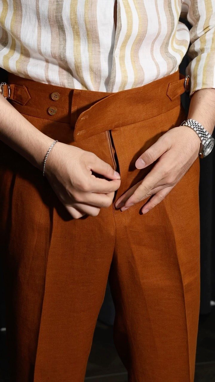 Rust-colored linen trousers with a high waist and button fly paired with striped shirt
