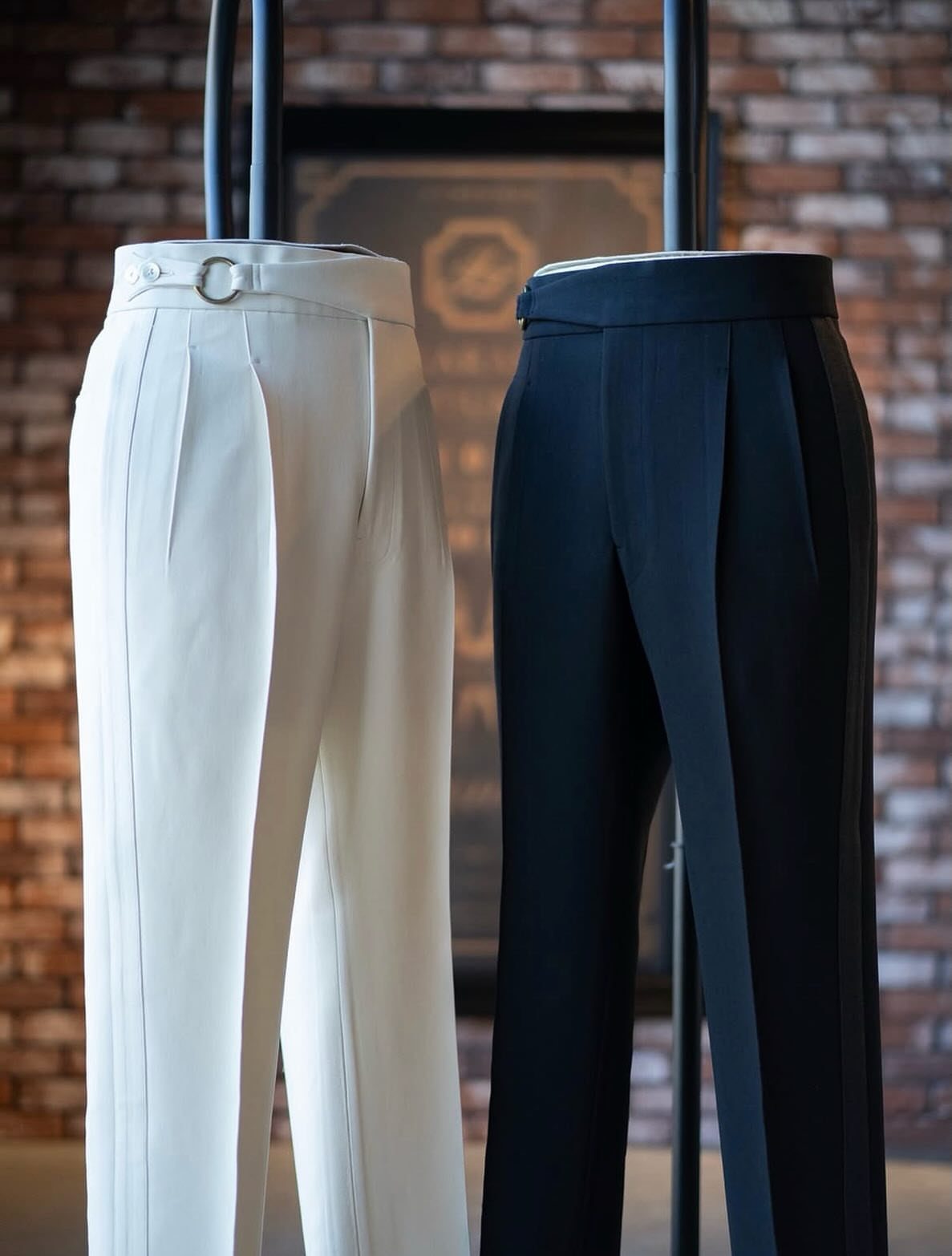 White and black pleated trousers hanging on a rack against a brick wall.