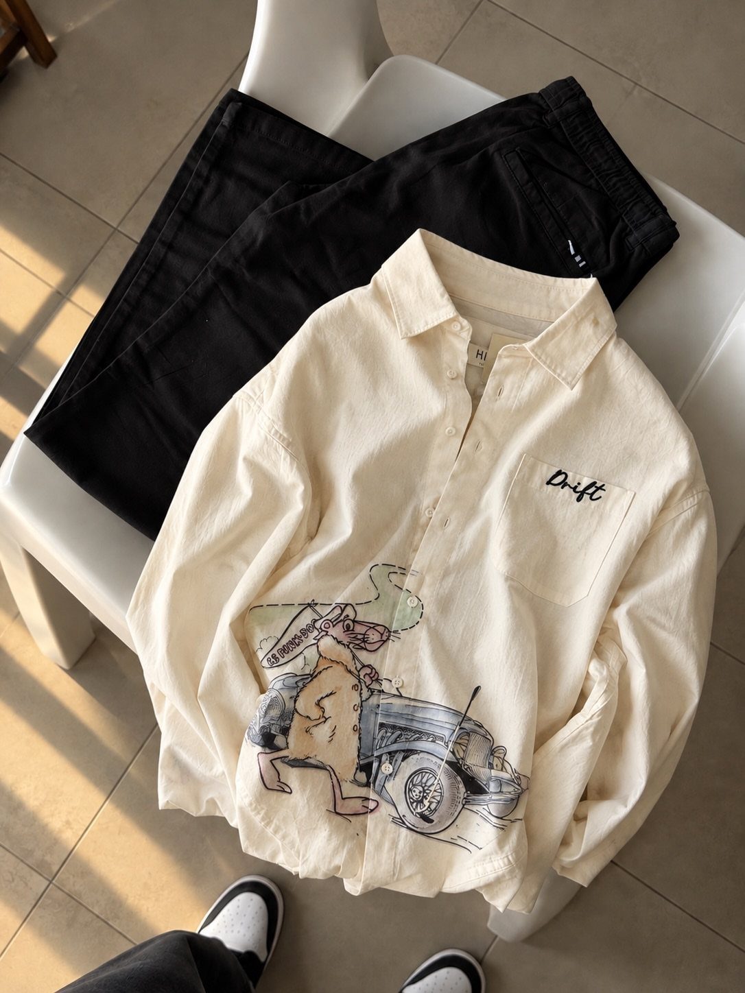 Cream long-sleeved shirt with vintage car graphic and black trousers on white chair