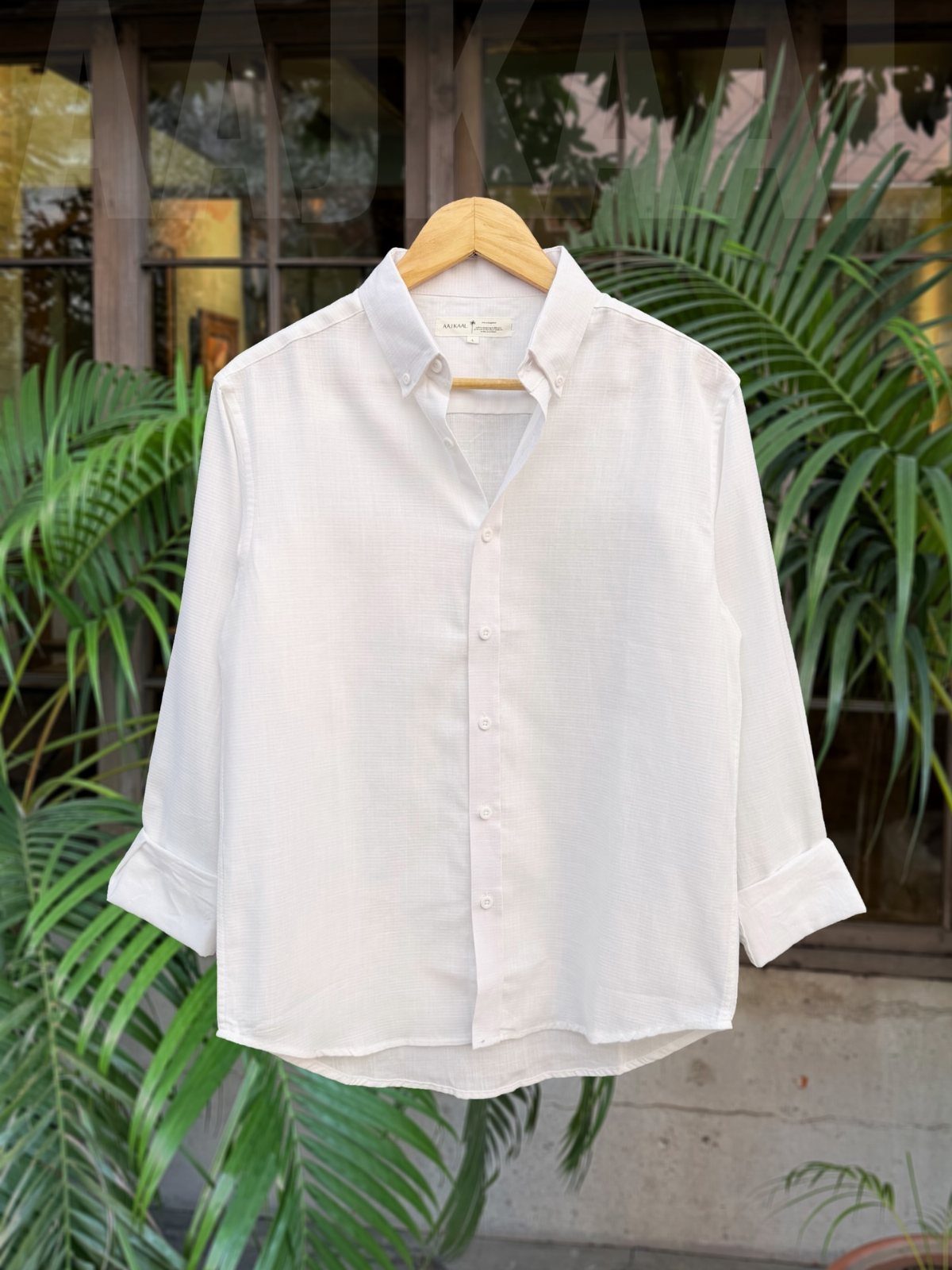 White button-down shirt hanging on a wooden hanger with green palm leaves behind.
