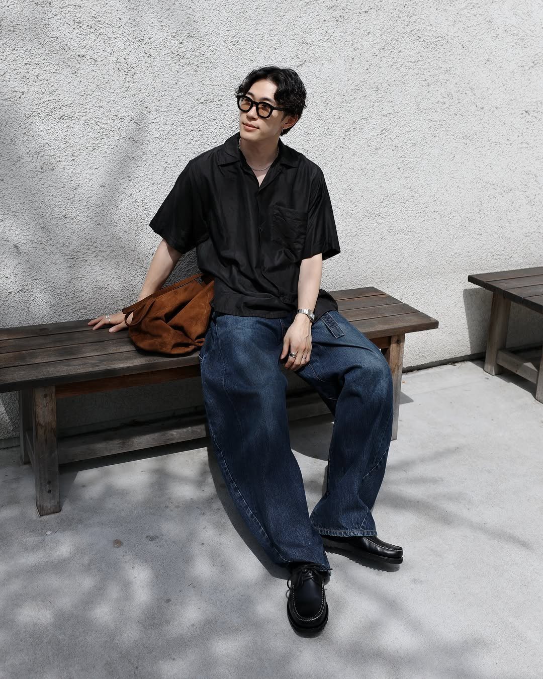 Man in black shirt and wide-leg jeans sitting on a wooden bench.
